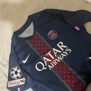 PSG matchtröja D. Doué #14 Nike - Snygg Paris Saint-Germain fotbollströja med D. Doué och nummer 14 på ryggen. Marinblå med rött och vitt geometriskt mönster längs mitten, PSG-logga på bröstet och Champions League-patch på ärmen. Tillverkad i lätt polyester, perfekt för match eller träning.