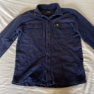 Mörkblå overshirt från Lyle & Scott - Snygg mörkblå overshirt från Lyle & Scott Junior med klassisk krage, två bröstfickor med lock och knappar framtill. Finns inga hål eller vidare på den, använd några få gånger