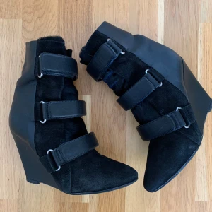 Svarta kilklackade stövletter med kardborre - Unika svarta stövletter med kilklack och tre breda kardborreband framtill. Skorna har en mix av mocka och skinn, spetsig tå och coola detaljer i metall. Perfekta för dig som vill sticka ut med en edgy och modern look.
