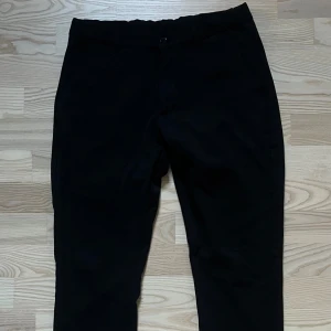 Svarta kostymbyxor från Grunt - Stilrena svarta kostymbyxor från Grunt med klassisk rak passform och hällor för bälte. Perfekta till en snygg skjorta eller stickad tröja. Byxorna har diskreta sidfickor och stängs med knapp och dragkedja.