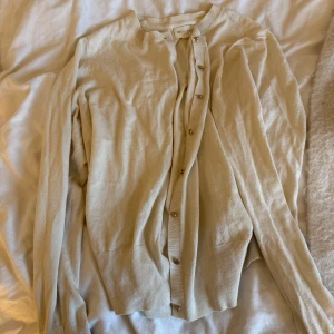 Beige kofta från Holly & Whyte - Snygg beige kofta från Holly & Whyte med rund halsringning och guldiga knappar framtill. Långärmad modell i mjukt material med ribbade muddar vid ärmslut och nederkant. Perfekt att slänga över en t-shirt eller linne för en chill look.