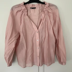 Ljusrosa blus med knappar från Zara - Supersöt ljusrosa blus från Zara med v-ringning och knappar framtill. Blusen har långa ärmar med lätt puff. Tunt och luftigt material. Behövs strykas. Aldrig använd