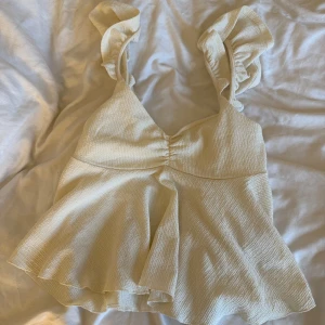 Krämvit volangig linnetopp - Supersöt krämvit linnetopp med volangiga axelband och lätt rynk framtill. Toppen har en v-ringad skärning och är sydd i ett strukturerat, lite stretchigt tyg som ger en lyxig känsla. Perfekt för varma dagar när du vill känna dig extra fin.