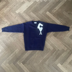 Mörkblå stickad tröja från Ami Paris - Snygg mörkblå stickad tröja från Ami Paris med klassisk rund hals och ribbade muddar. På bröstet finns Ami:s ikoniska hjärta med bokstaven A i vitt. Perfekt för dig som gillar stilrena och trendiga plagg med en twist.
