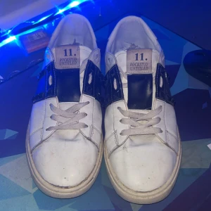 Valentino Rockstud Untitled sneakers - Snygga vita Valentino Rockstud Untitled sneakers med svarta detaljer och ikoniska nitar på sidorna. Skorna har snörning, platt sula och rund tå. Tillverkade i skinn för en lyxig känsla och streetig look. Perfekta för dig som vill sticka ut med designer vibes.