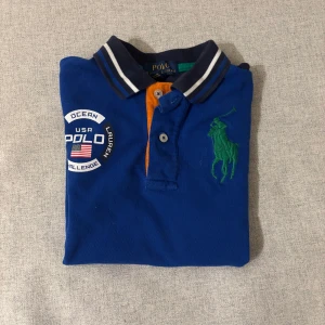 Blå Polo Ralph Lauren t-shirt - Storlek s eller 152                                                 Snygg blå t-shirt från Polo Ralph Lauren med korta ärmar och vit panel på sidorna. Stor grön polospelare broderad på bröstet, patch med USA-flagga och Polo Ocean Challenge på ena sidan. Kragen har marinblå och vita ränder samt orange knappslå. Perfekt för dig som gillar sportig stil.