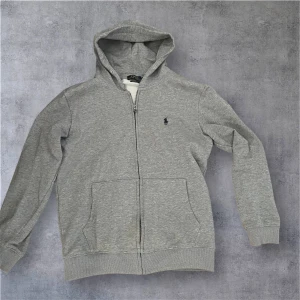 Grå Kofta från Polo Ralph Lauren - Storlek passar båda M och S Snygg grå hoodie från Polo Ralph Lauren med dragkedja och klassisk broderad logga på bröstet. Tröjan har huva med snörning, två stora fickor fram och ribbade muddar. Perfekt för en chill och stilren look.