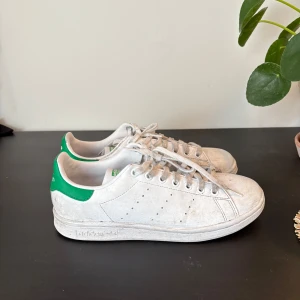 Adidas Stan Smith vita sneakers - Klassiska Adidas Stan Smith sneakers i vitt med gröna detaljer på hälen och tungan. Skorna har snörning, rund tå och platt sula. Perfekta för en clean och tidlös stil. Tillverkade i syntetmaterial med diskreta perforeringar på sidorna.