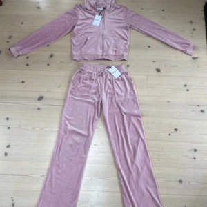 Rosa luvjacka och byxor från Juicy Couture - Mysiga rosa mjukisbyxor från Juicy Couture i mjuk velour med broderad JC-logga på fickan. Byxorna har dragsko i midjan, raka ben och stora fickor både fram och bak. Perfekta för en chill och trendig look. Jacka med dragkedja. Nypris 899:-/del säljer nu båda för 1100:-