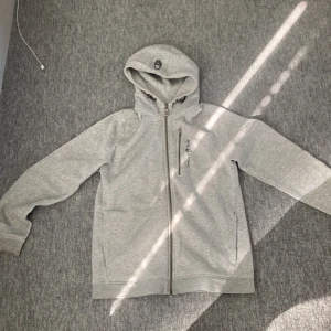 Ljusgrå hoodie från Sail Racing - Ljusgrå hoodie från Sail Racing med dragkedja och huva. Tröjan har broderad logga på bröstet och är helfodrad i mjuk fleece. Använd och har en väldigt liten fläck 