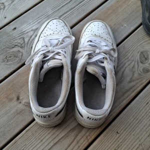 Nike Air Force 1 vita sneakers - Klassiska Nike Air Force 1 sneakers i vitt skinn med svart Nike-logga på hälen. Skorna har snörning, perforerad tå och platt sula. Perfekta för dig som gillar en clean och tidlös stil. Snygga till jeans eller shorts.