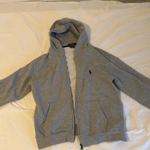 Grå hoodie från Polo Ralph Lauren - STORLEK M fast passar S ! Snygg grå hoodie från Polo Ralph Lauren med dragkedja och huva. Klassisk broderad logga på bröstet och två fickor framtill. Mjuk bomullskvalitet och normal passform, perfekt för chill dagar. Den är använd 1-5 gånger säljs pvg storlek