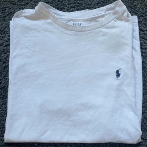 Vit t-shirt från Polo Ralph Lauren - Vit t-shirt från Polo Ralph Lauren med klassisk rund hals och korta ärmar. T-shirten har den ikoniska lilla broderade Polo-loggan i mörkblått på bröstet. Tillverkad i mjuk bomull som känns skön mot huden. Perfekt för en clean och stilren look.