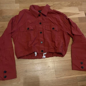 Röd croppad overshirt från Lindex - Croppad röd overshirt från Lindex med knappar framtill, två bröstfickor och dragsko i midjan. Jackan har klassisk krage och långa ärmar med knappdetaljer vid ärmslut.