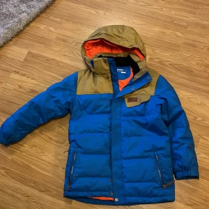Blå och brun dunjacka från Everest - Säljer en blå och brun dunjacka från Everest med orange foder. Jackan har huva, dragkedjor på fickorna och tryckknappar framtill. Perfekt för kalla dagar och har en sportig look med snygga färgblock.