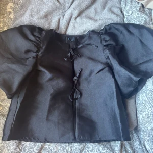 Svart blus med puffärmar från Gina Tricot - Supersnygg svart blus från Gina Tricot i storlek XS. Blusen har stora puffärmar och knytband framtill som ger en trendig look. Perfekt för dig som vill sticka ut med en cool och unik topp. Materialet känns slätt och lätt, och modellen är lite croppad.