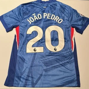 Chelsea João Pedro #20 matchtröja Nike - Chelsea FC fotbollströja med João Pedro och nummer 20 på ryggen. Tröjan är blå med diskret grafiskt mönster, vita detaljer och klubbmärke på bröstet. Tillverkad i lätt och ventilerande Dri-FIT-material. Premier League-logga på ärmen. Replika, endast handtvätt gäller!