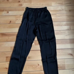 Svarta satinbyxor från ARKET, strl 34 - Svarta satinbyxor från ARKET i storlek 34. Byxorna har elastisk midja med dragsko och är tillverkade i ett glansigt, mjukt material som känns riktigt skönt mot huden. 
