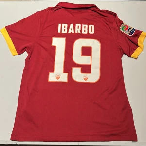 AS Roma Ibarbo 19 matchtröja Nike - AS Roma fotbollströja med Ibarbo och nummer 19 på ryggen. Röd med gula detaljer, officiellt Serie A-märke på ärmen och klubbmärke på bröstet. Tillverkad i Dri-FIT-material för bästa komfort. Klassisk pikékrage och korta ärmar. Replika, endast handtvätt gäller!