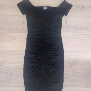 Svart omlottklänning med rynk - Snygg svart kortklänning med offshoulder och rynkade detaljer längs hela framsidan. Klänningen har en tight passform och är tillverkad i ett mjukt, stretchigt material som sitter skönt mot kroppen. Perfekt för dig som gillar en stilren look med lite edge.