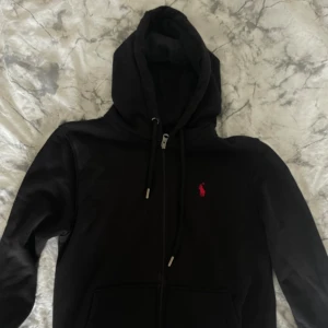 Svart hoodie från Polo Ralph Lauren - Svart hoodie från Polo Ralph Lauren med röd broderad logga på bröstet. Tröjan har huva med snörning, dragkedja framtill och två stora fickor. Materialet är mjuk bomull och passformen är normal. Perfekt för chill dagar eller när du vill ha en clean look.