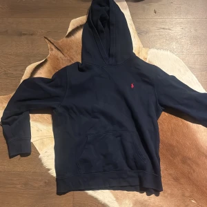 Mörkblå hoodie från Polo Ralph Lauren - Snygg mörkblå hoodie från Polo Ralph Lauren med klassisk röd broderad logga på bröstet. Tröjan har huva, känguruficka och långa ärmar. Perfekt för dig som gillar stilren och bekväm streetwear.