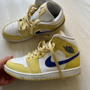 Nike Air Jordan 1 gul och vit - Nike Air Jordan 1 sneakers i en fräsch kombo av vitt och ljusgult skinn med blå swoosh och Air Jordan-logga på sidan. Klassisk high-top modell med snörning och perforerad tå för extra vibe. Perfekta för dig som vill sticka ut med färg på fötterna.