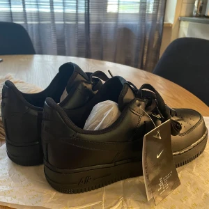 Nike Air Force 1 helsvarta  - Säljer ett par klassiska Nike Air Force 1 Low i helsvart färg. Skorna har en snygg, stilren design med rund tå, platt sula och snörning. Tillverkade i skinn med perforerade detaljer på tån för extra ventilation. Perfekt för dig som gillar streetstyle och vill ha en ikonisk sneaker.