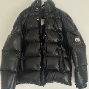 Svart Moncler Maya 70th Anniversary - Säljer en svart pufferjacka från Moncler med glansig finish och ikonisk patch på ärmen. Jackan har dragkedja och tryckknappar framtill samt luva och praktiska fickor. Insidan har ett coolt serietryck. Perfekt för dig som vill ha en varm och snygg vinterstil.