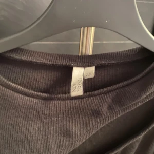 Svart ribbad långärmad topp NY Trend - Svart långärmad topp från NY Trend i ribbat material. Toppen har en unik asymmetrisk cut out-detalj över bröstet som ger en cool och modern look. Passformen är slim och tyget är stretchigt för extra komfort. Perfekt för dig som vill sticka ut lite extra.