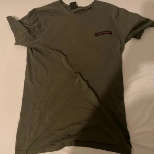 Olivgrön t-shirt från Hugo Boss - Snygg olivgrön t-shirt från Hugo Boss med diskret logga i rött och svart på bröstet. Klassisk rund halsringning och korta ärmar. Skön och mjuk känsla, perfekt till jeans eller shorts. Stilren design som funkar året runt.