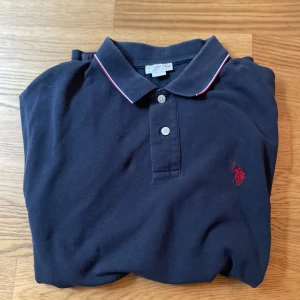 Svart pikétröja från U.S. Polo Assn. - Snygg marinblå pikétröja från U.S. Polo Assn. med klassisk krage, två knappar och röd logga broderad på bröstet. Tunn röd och vit kant på kragen och ärmsluten ger en clean look. Tillverkad i mjuk bomull, perfekt för en stilren vardagsoutfit.