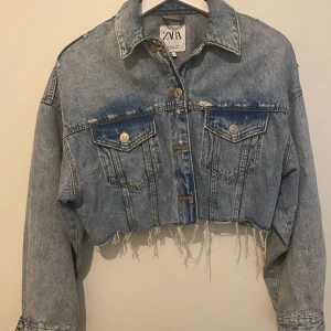 Croppad jeansjacka från Zara XS - Säljer en croppad jeansjacka från Zara i ljusblå denim. Jackan har rå, fransig nederkant, två bröstfickor med knappar och slitna detaljer framtill. Klassisk krage och långa ärmar. Perfekt för dig som gillar streetstyle och edgy vibe.