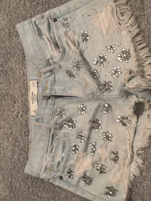 Ljusa jeansshorts med paljetter från Abercrombie & fitch  - Pris kan diskuteras Säljer ett par ljusblå jeansshorts från Abercrombie & Fitch med slitna detaljer, fransig kant och blommiga paljetter framtill. Shortsen har normal passform och är perfekta för sommarens alla äventyr. Klassisk femficksmodell och tillverkade i bomull.