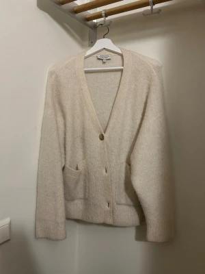 Vit kofta från & Other Stories - Mysig beige kofta från & Other Stories Los Angeles Atelier. Koftan är ribbstickad med v-ringning, två framfickor och knappar framtill. Perfekt för lager på lager och har en relaxed, lite loose passform.Ursäkta dålig belysning. 