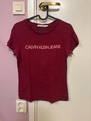 Vinröd t-shirt Calvin Klein Jeans - Snygg vinröd t-shirt från Calvin Klein Jeans med klassisk logga tryckt i vitt över bröstet. Modellen är kortärmad och har en rund halsringning. Tillverkad i mjuk bomull som känns skön mot huden. Perfekt till jeans eller kjol för en clean och enkel look.