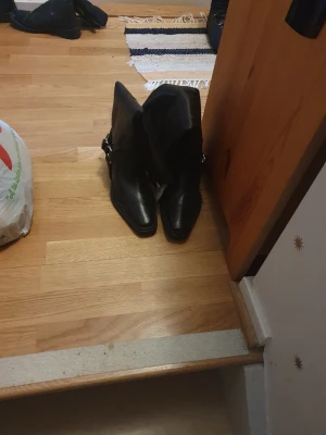 Svarta cowboy boots med spänne - Säljer ett par svarta cowboy boots i skinn med coolt spänne på sidan. Bootsens tå är spetsig och sulan är platt, vilket ger en edgy look. Perfekta för dig som vill sticka ut och addera lite western-vibe till outfiten.