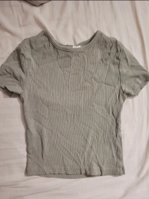 Ljusgrå ribbad kortärmad topp - En enkel och stilren ljusgrå topp med ribbad struktur. Modellen är croppad och har korta ärmar samt rund halsringning. Perfekt att matcha med jeans eller kjol för en avslappnad look.