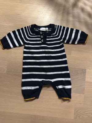 Randig body från Name It, marinblå/vit - Säljer en söt randig body från Name It i marinblått och vitt. Bodyn har långa ärmar, knappar framtill och tryckknappar i grenen för enkel påklädning. Ribbad hals och muddar med en gul detalj vid ärmslut och benslut. Perfekt för små bebisar!