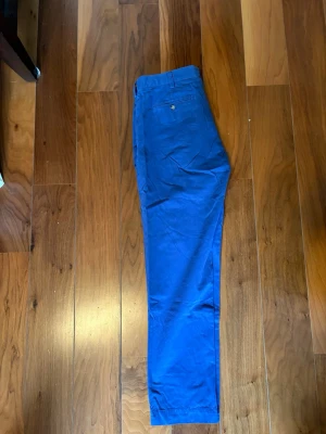 Blå chinos från Polo Ralph Lauren - Snygga blå chinos från Polo Ralph Lauren i slim fit-modell. Byxorna har klassisk design med bakficka med knapp och är tillverkade i mjukt bomullstyg. Perfekta för dig som gillar stilrena och bekväma byxor.