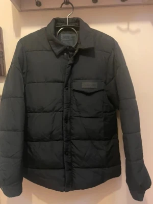 Svart pufferjacka - Säljer en svart pufferjacka från med klassisk krage och knäppning framtill. Jackan har en bröstficka med lock och diskret logga, samt quiltad design som ger en schysst look. Perfekt för kyliga dagar och enkel att matcha med allt. Fint skick, knappt använd.