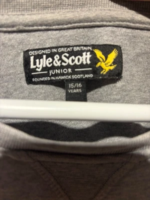 Grå sweatshirt från Lyle & Scott  - Klassisk grå sweatshirt från Lyle & Scott Junior med rund halsringning och den ikoniska gula logon broderad på bröstet. Tröjan har långa ärmar och ribbade muddar vid ärmslut och nederkant. Perfekt för en avslappnad och stilren look.