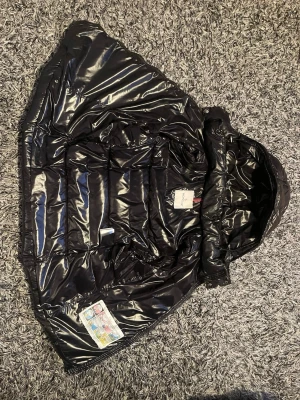 Moncler väst med luva  - Säljer en svart, glansig dunväst från Moncler med huva och dragkedja framtill. Västen har två fickor med dragkedja och ett klassiskt Moncler-märke på bröstet. Perfekt för lager-på-lager och riktigt snygg till höst och vinter.