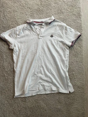 Vit Moncler piké med rödblå detaljer - Säljer en klassisk vit pikétröja från Moncler i storlek L. Pikén har korta ärmar, krage med röd och blå rand samt Moncler-logga broderad på bröstet. Materialet är mjuk bomull och modellen har knappar vid halsen. Perfekt för en clean och sportig look.