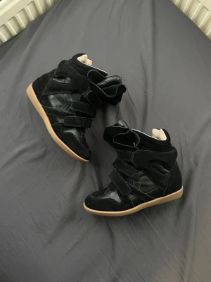 Isabel Marant svarta sneakers med kilklack - Säljer ett par svarta Isabel Marant sneakers med kilklack och kardborreband. Skorna har en mix av mocka och skinn, beige insida och gummisula i naturfärg. De har en chunky siluett och högt skaft som ger en riktigt cool streetstyle-vibe.