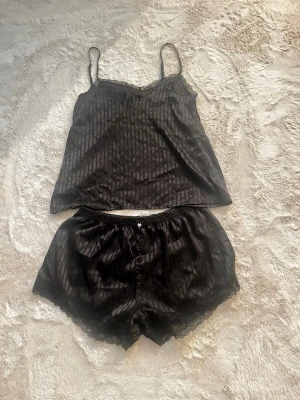 Svart randig Satin Pyjamas  - Säljer en svart pyjamas i satin från New Yorker med tunna axelband och randigt mönster. Överdelen har spetsdetaljer vid urringningen och en mjuk, glansig finish som ger en lyxig känsla.