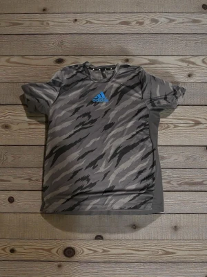 Grå camo t-shirt från Adidas - Säljer en grå t-shirt från Adidas med camo-inspirerat mönster i olika grå toner och svart. Framsidan har en blå Adidas-logga och baksidan är mestadels enfärgad. Tillverkad i lätt polyester, perfekt för träning eller chill. Klassisk passform och korta ärmar.