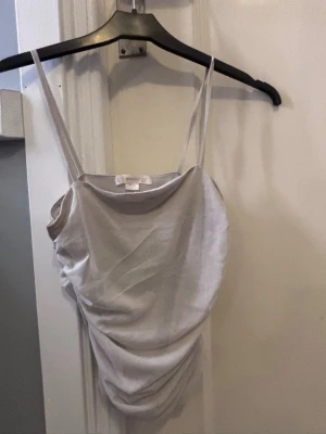 Beige linnetopp från Gina Tricot - Säljer en beige linnetopp från Gina Tricot med tunna axelband och snygg rynkning på sidan. Toppen har en mjuk, lätt glansig yta och är perfekt för varma dagar. Enkel och stilren design som är lätt att matcha.