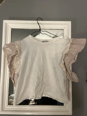Vit topp med volangärmar från Zara - Supersöt vit topp från Zara med stora, vågiga volangärmar i beige. Toppen har rund halsringning och är gjord i mjuk bomull. Perfekt för dig som vill ha något extra till din outfit. Volangdetaljerna ger en unik och trendig look. Passar de som har S också.