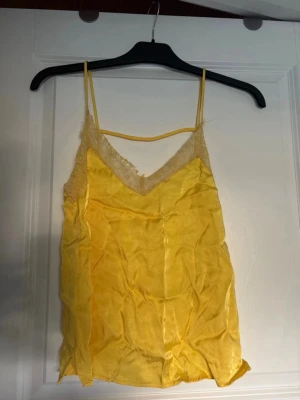 Gul linnetopp med spetsdetaljer - Säljer en solgul linnetopp med tunna axelband och v-ringning. Toppen har fina spetsdetaljer längs ringningen och är gjord i ett glansigt, mjukt material som ger en lyxig känsla. Perfekt för varma dagar eller att styla lager på lager.